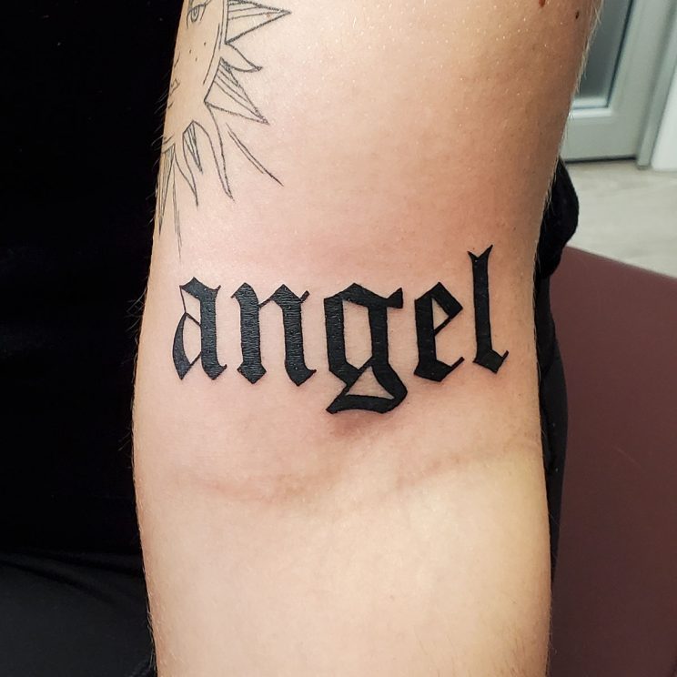 Old English style script of the word "Angel" just below bicep.